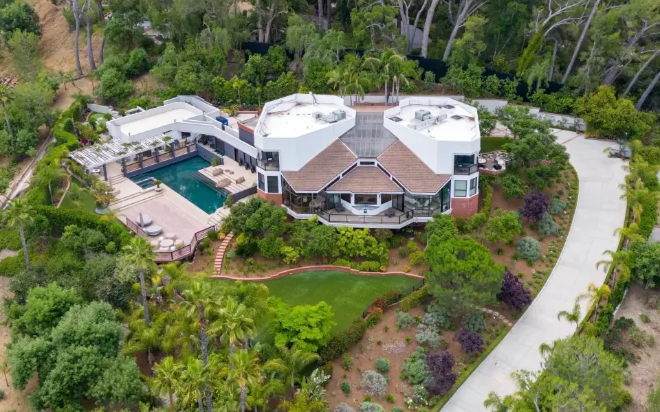 Aerial view of a large, Pat Sajak's stunning Encino House surrounded by trees and greenery with a long driveway leading to the property and a swimming pool in the backyard.