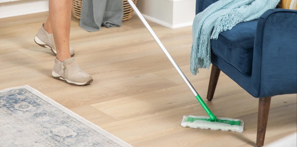 Person using a Swiffer Sweeper with a dry cloth to clean laminate flooring next to a blue chair and rug.