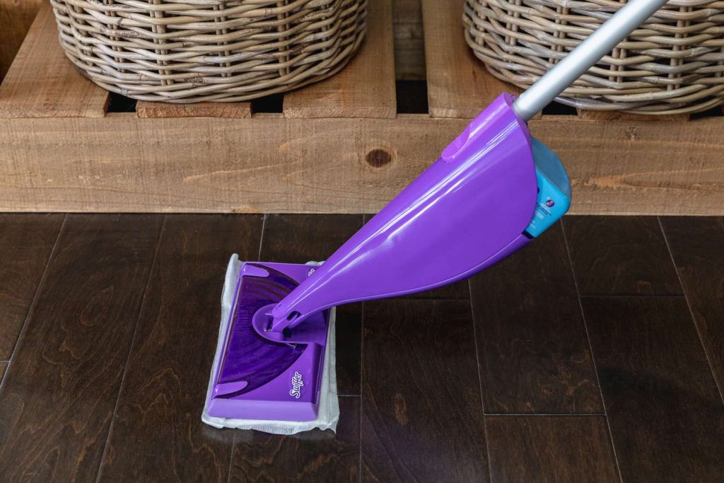 Close-up of a purple Swiffer WetJet mop with a cleaning pad attached, being used on dark hardwood flooring.