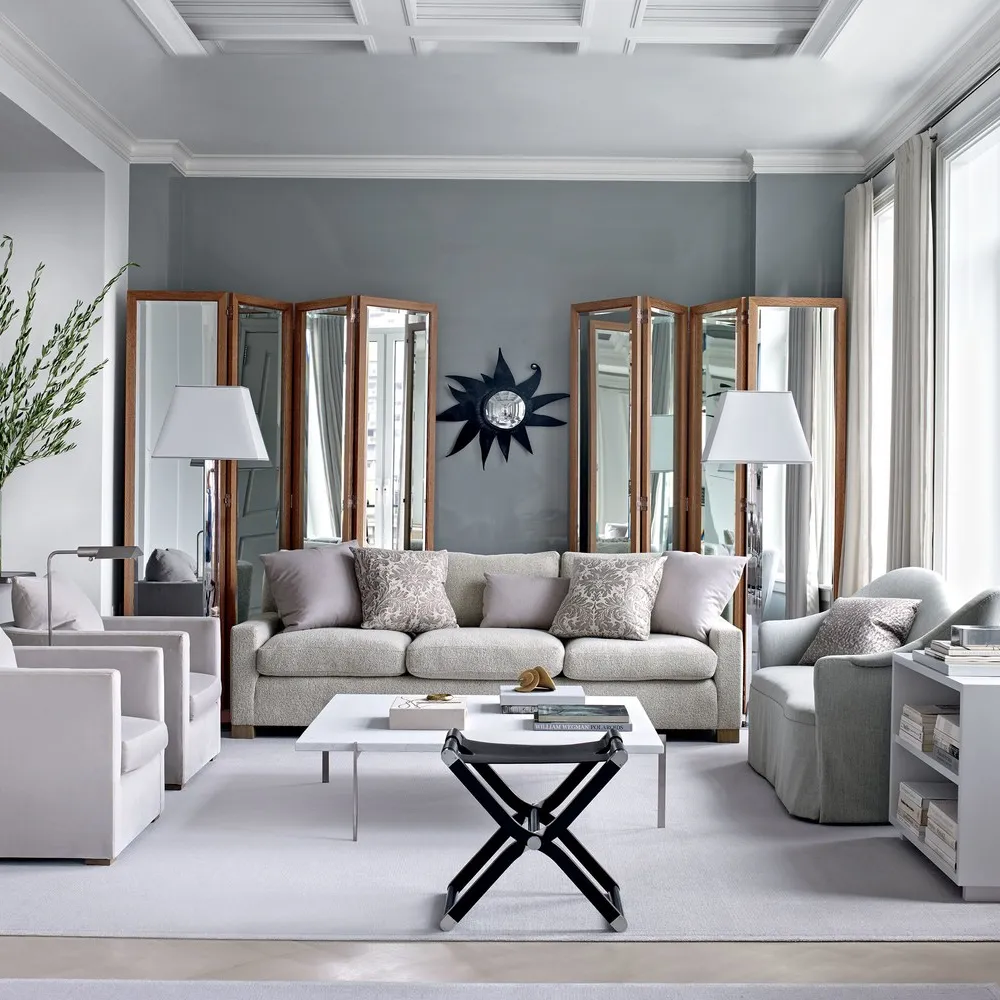 Elegant living room with cool gray walls, light neutral sofas and armchairs, mirrored panels, modern coffee table, and soft natural light from tall windows.