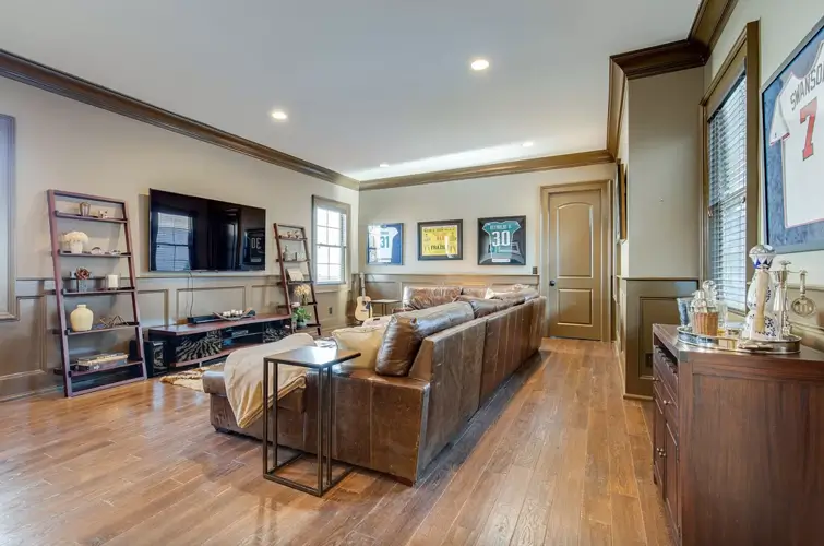 The interior of Theo Von's Nashville home, showing a cozy family room or den with dark hardwood floors, a large brown leather L-shaped sofa, framed sports jerseys on the wall, and a TV stand.