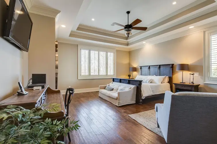 The master bedroom of Theo Von's Nashville home, featuring dark hardwood floors, a large dark wood bed frame, a white sofa at the foot of the bed, and a wooden desk area.