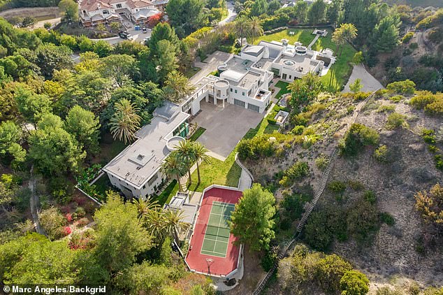 Aerial view of a luxury mansion surrounded by lush greenery featuring modern architecture, spacious driveway, and a red tennis court among landscaped grounds
