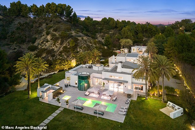 Aerial view of a luxury modern mansion showcasing its outdoor patio area with a glowing pool, sleek white architecture, circular glass tower, and palm trees at dusk