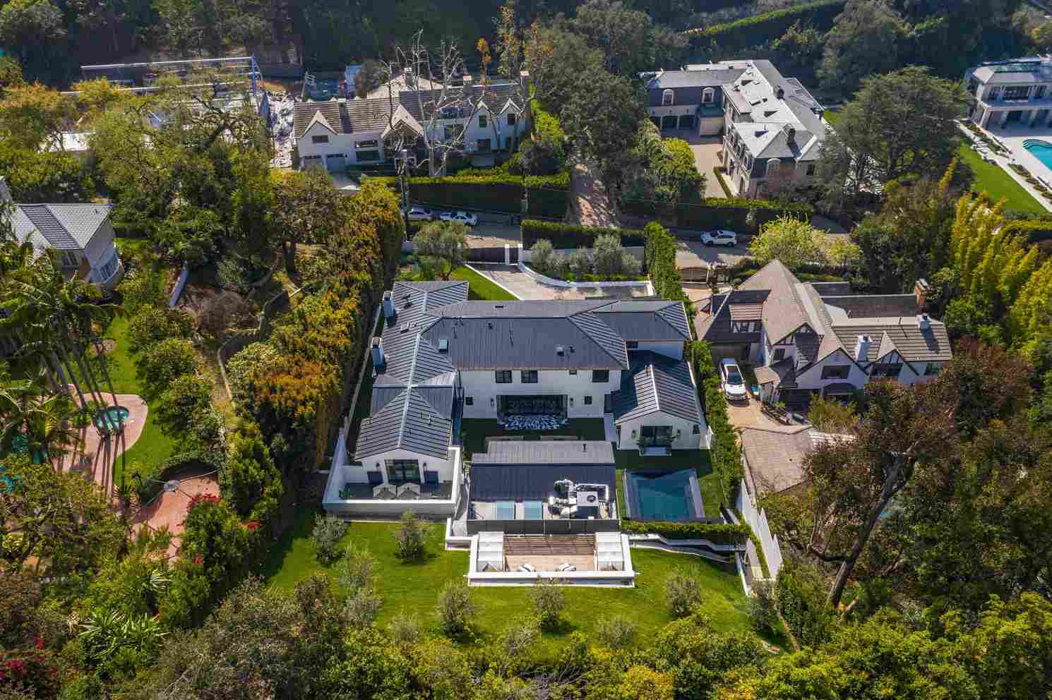 Aerial view of a sprawling modern luxury mansion in Beverly Hills, featuring white architecture, dark roof, manicured lawns, swimming pool, and surrounded by lush greenery and neighboring homes.