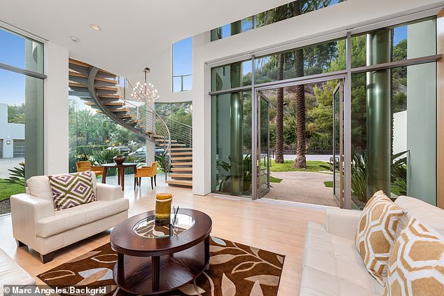 Bright modern living room in a luxury mansion featuring a spiral staircase, floor-to-ceiling glass walls opening to an outdoor garden, and light wood flooring