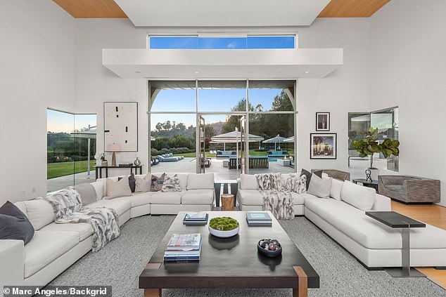 Bright modern living room with a large L-shaped white sectional sofa, dark coffee table, and expansive glass windows overlooking an outdoor patio