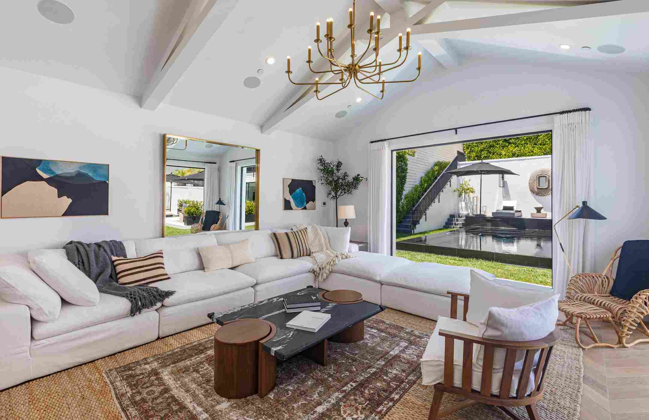 Elegant Beverly Hills living room featuring white sectional sofa, gold chandelier, abstract art, and floor-to-ceiling windows revealing outdoor pool and garden area with contemporary design elements.