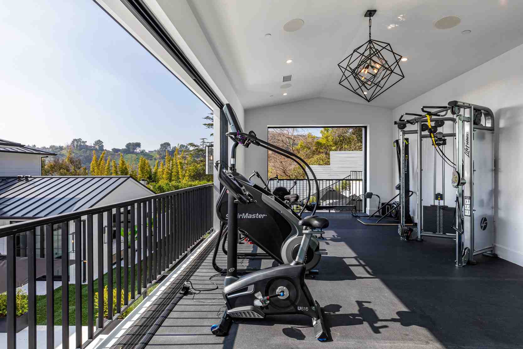 Luxury home gym in a Beverly Hills mansion featuring high-end exercise machines, geometric lighting, and expansive windows showcasing scenic hills and neighboring homes.
