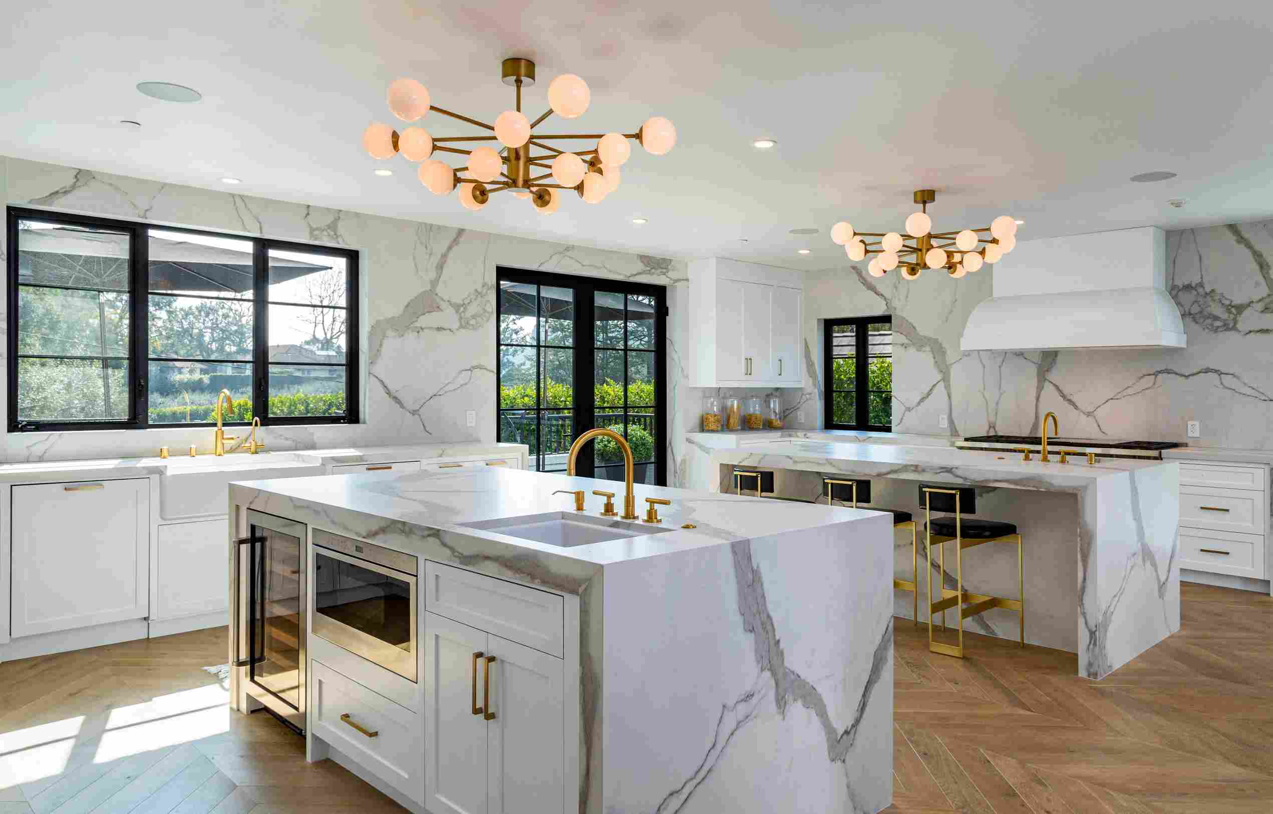 Spacious modern kitchen in a Beverly Hills mansion featuring marble countertops, gold fixtures, white cabinetry, and large windows overlooking greenery, illuminated by stylish chandeliers.