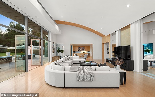 Spacious sunlit luxury living room with a curved white sectional sofa, curved wooden ceiling, floor-to-ceiling glass windows, and sleek black entertainment unit