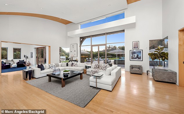Spacious sunlit luxury living room with high ceilings, floor-to-ceiling windows, curved wooden ceiling detail, light wood flooring, and plush white sectional sofa