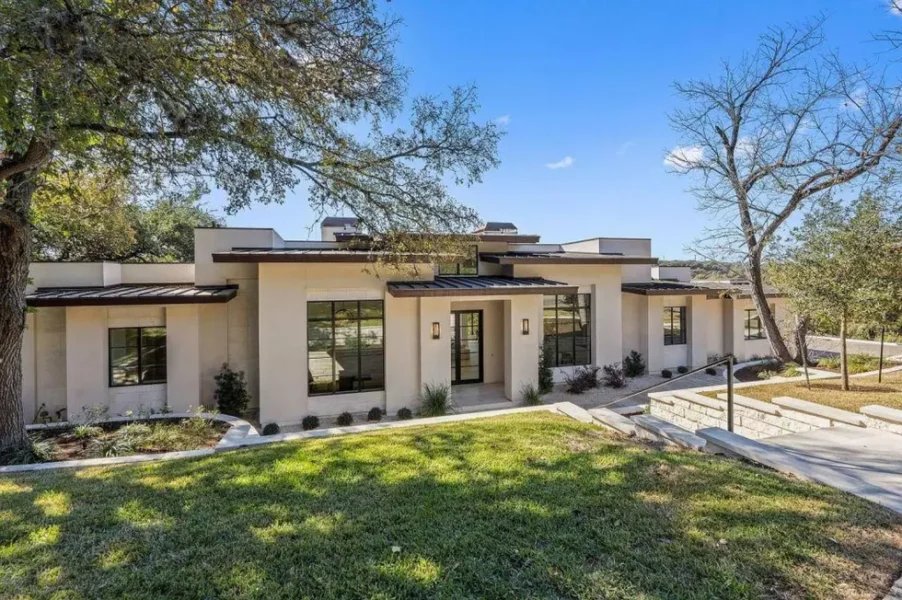 Tom Segura’s modern Austin house on Lake Austin: light stucco exterior, metal roof, large windows, and landscaped lawn.